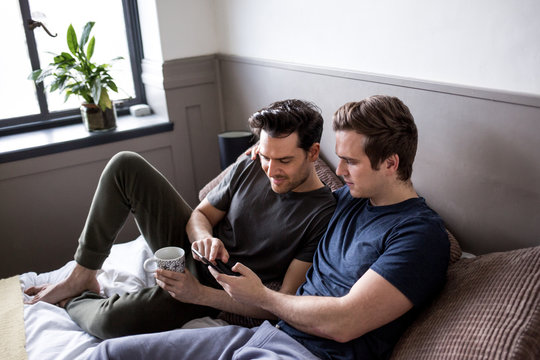 Gay couple using a mobile phone while leaning on bed