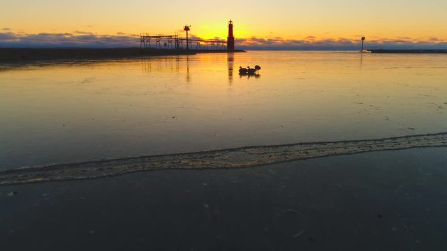 Three hearty cold weather geese walk on thin ice before sunrise, Iconic Lighthouse silhouetted against fiery Lake Michigan twilight, sunrise.