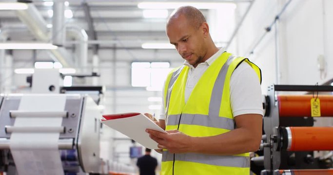 4K Worker in printing factory looking down at paperwork with machinery in background