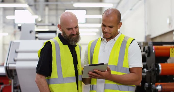 4K Coworkers in a printing factory looking down at computer tablet, with machinery in background