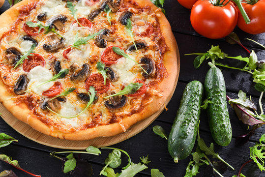 Delicious homemade pizza with mushrooms, tomatoes, mozarella and arugula served with fresh vegetables and herbs on black background