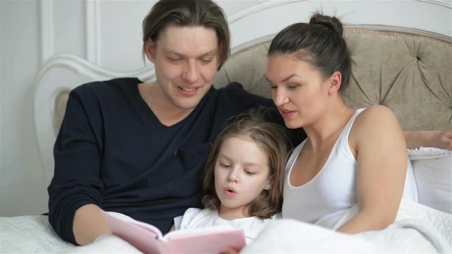 Clever Little Girl is Reading Aloud a Book Lying on the Bed. Her Young Parents are Helping Her. Female Child is Learning Foreing Language with Family.