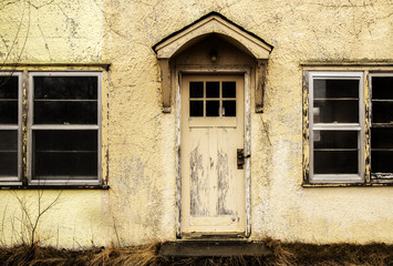 A close up of a cracked and faded exterior cement wall of a home with a door with wood awning above and a window on both sides