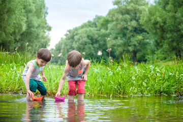 Two little brothers playing with paper boats by a river on warm and sunny summer day.