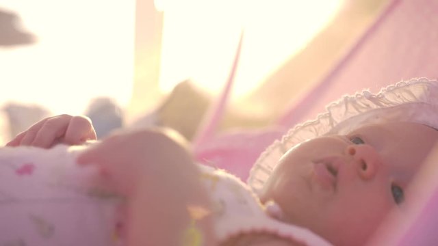 Slow motion close-up shot of three months baby girl in bonnet lying in pink bassinet outdoor in summer. View in warm evening sunlight