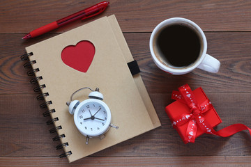 Notebook with heart, the alarm clock, a cup of coffee and a gift on a table