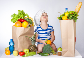 Happy little girl in the hat of the chef with big bags of food. A variety of fresh fruits and...