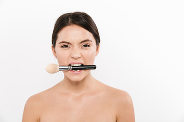 Studio portrait of cheerful asian woman being half-naked and makeup brush, in her mouth, isolated over white background