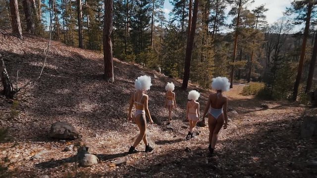 Extravagant women dressed in white underwear and wigs are going in sunny summer woodland. Back view of their slim and graceful bodies