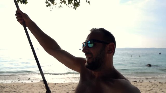 Happy young man taking selfie, video during swinging on the beach, slow motion
