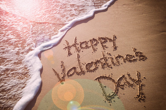 Happy Valentine's Day message handwritten on smooth sand beach with incoming wave and romantic lens flare