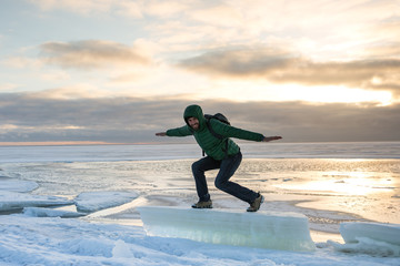 Fototapeta na wymiar Joyful bearded man in green jacket, jeans standing, balances and surfing on ice floe in winter day/ Man stands on an ice floe against the frozen sea, cloudy sky on background