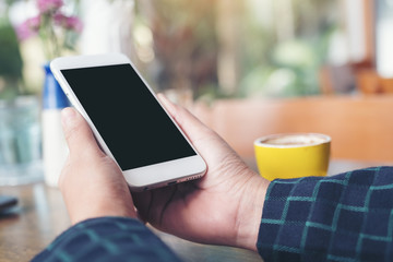 Mockup image of a hand holding white mobile phone with blank black desktop screen and yellow coffee cup on wooden table in modern cafe