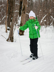 The girl in the green suit walk slowly through the woods on skis.