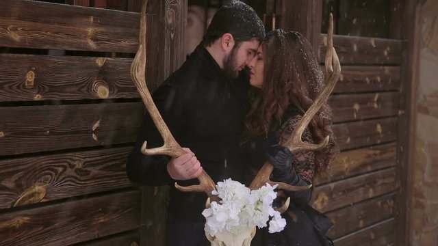 A loving couple in black clothes stands embracing and holding a skull of a deer in their hands, they stand outdoors in case of a heavy snowfall against the background of a wooden fence.