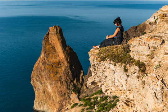 Beautiful girl sitting on a high rock and looking out to sea. Girl sits on the edge of cliff. The girl of the sea. Seascape. Girl at sunset