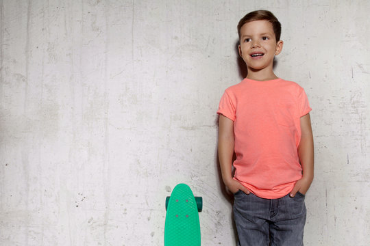 Cute little guy posing with skateboard against gray wall. Boy with skateboard outdoors standing near gray wall, copy space. Portrait of skater boy in pink T-shirt, concrete wall on background.