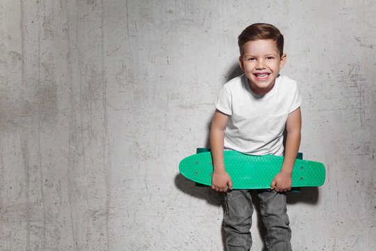 Attractive little boy with green skateboard in his hands. Smiling guy in white T-shirt standing near grunge concrete wall. Concept of activity and happy childhood.