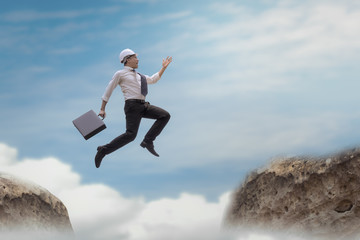 Businessman with helmet jumping over gap on the rock with sky background, obstacle and problem gap concept