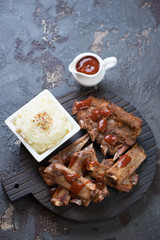 Black wooden serving board with baked pork ribs, potato puree and sauce. High angle view on a brown stone background