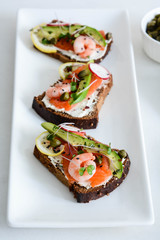 Smorrebrod, traditional Danish open sanwiches, dark rye bread with salmon, avocado, shrimp and micro-greenon a white plate. 
