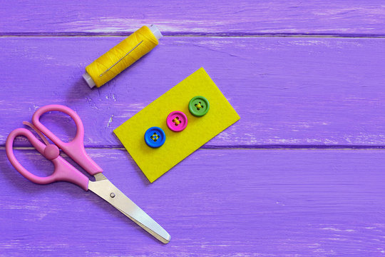 How to sew on a button with four holes. Colored buttons on yellow felt piece. Scissors, thread, needle on a wooden background with copy space for text. Wooden button decoration idea. Top view 