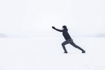 Fototapeta na wymiar Young man in dark sporty wear alone doing exercise before running at white snow covered place. People's daily outdoor active lifestyle in cold winter season. Enjoying sport and freedom concept.