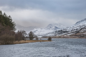 Beautiful Winter landscape image of Llynnau Mymbyr in Snowdonia National Park with snow capped mountains in background