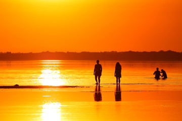 Silhouettes of people in shallow water in the lake with healing mud at orange red sunset