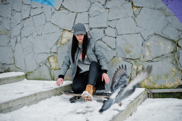 Stylish brunette girl in gray cap, casual street style with skate board on winter day against colored wall.