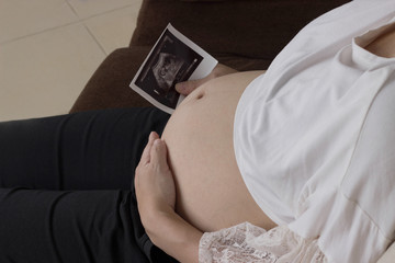 Close up of  pregnant woman holding x-ray photo of her baby near big belly. She is sitting on sofa .