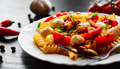 fusilli pasta salad with tomato, pepper and mozzarella balls in plate on dark wooden background