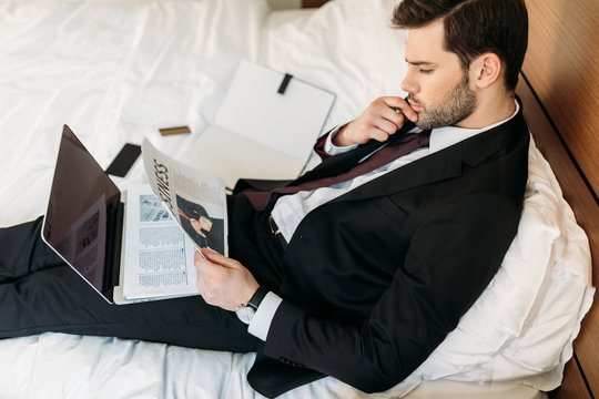 high angle view of businessman lying on bed in hotel room and reading newspaper