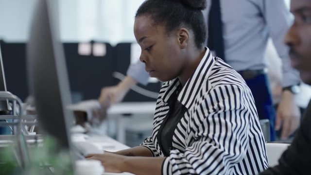 Tracking shot of concentrated African business lady in striped shirt typing on computer while working with colleagues in open space office