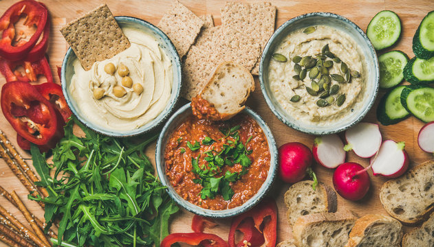 Vegan snack board. Flat-lay of various Vegetarian dips hummus, babaganush and muhammara with crackers, bread and fresh vegetables, wooden background, top view. Clean eating, dieting food concept