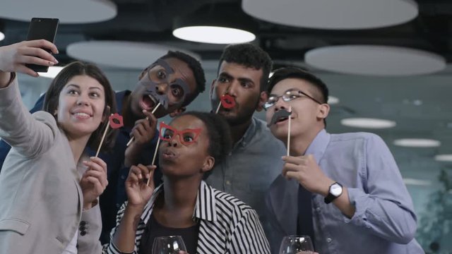 Young multi-ethnic team of business men and women holding funny mustache and eyeglasses on sticks, smiling and grimacing at camera of smartphone while taking picture at office party