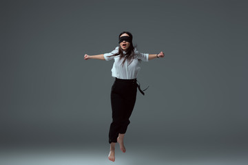 young barefoot woman in black blindfold jumping with open mouth on grey