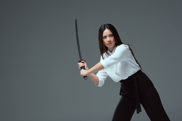 young asian woman fighting with katana sword and looking at camera isolated on grey