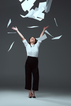 full length view of happy young businesswoman throwing papers on grey
