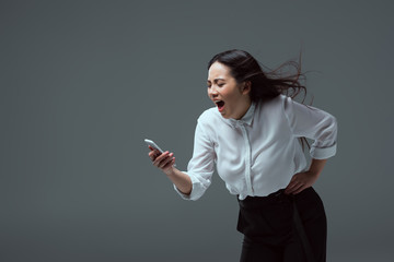 emotional asian girl holding smartphone and yelling isolated on grey