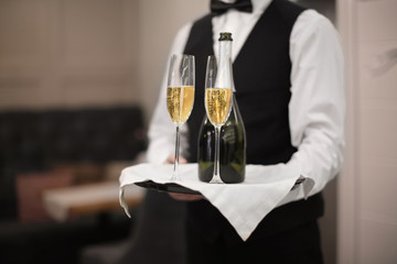 Waiter holding tray with glasses and bottle of champagne indoors, closeup