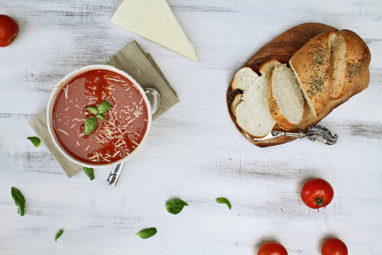 Hot tomato soup with basil leaves, parmesan cheese, fresh tomatoes and a loaf of herbed crusty bread. Image shot from above in flat lay style.