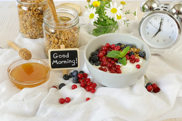 A bowl with yogurt, granola, blueberries and cranberries, granola jar, honey and flowers, alarm clock, good morning and healthy breakfast concept.