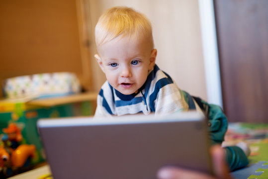Adorable blue eyed baby kid sitting on the bad and looking on a tablet at home.