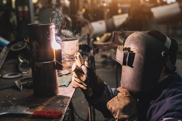 Welder in protective uniform and mask welding metal pipe on the industrial table while sparks flying.
