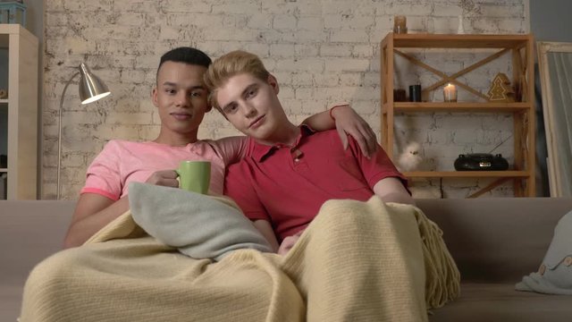 Two gay guys sit on the couch and watch TV, drink hot tea, use the remote control. LGBT lovers, a multinational couple, a happy gay family, a home cosiness concept. Look at the camera 60 fps