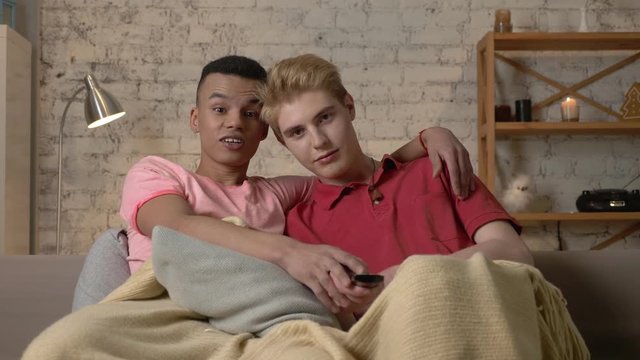 Two gay guys are sitting on the couch and watching TV, a Caucasian guy wants to switch the channel but an African guy stops him. LGBT lovers, a multinational couple, a happy gay family, a home