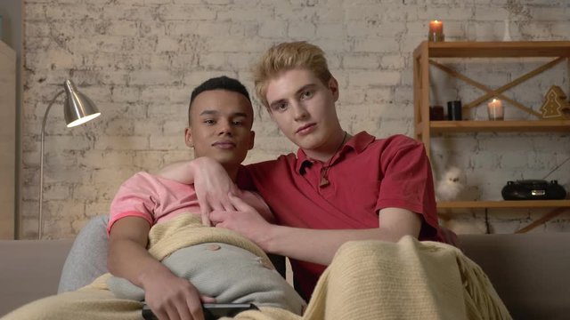 Two gay guys are sitting on the couch and watching TV, an African guy wants to switch the channel but an American guy stops him. LGBT lovers, a multinational couple, a happy gay family, a home