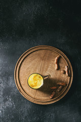 Cup of ayurvedic drink golden milk turmeric latte with curcuma powder on round wooden tray and ingredients above over black texture background. Top view, space. Toned image