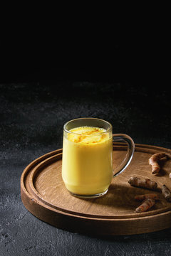 Cup of ayurvedic drink golden milk turmeric latte with curcuma powder on round wooden tray and ingredients above over black texture background. Copy space